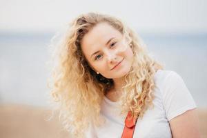 Outdoor shot of curly beautiful young female with fresh skin, looks positively at camera, wears casual white t shirt, poses against blurred seaside background. People and recreation concept. photo