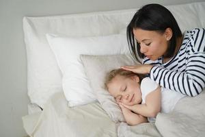 la madre pone a la hija en la cama, acostada sobre una almohada suave. siesta diurna, sueño saludable en niños, cuidado de niños foto