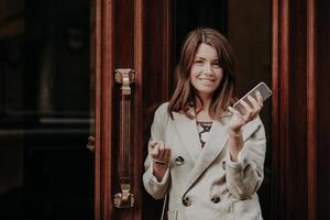 Elegant gorgeous lady in raincoat, holds mobile phone, waits for call, poses outdoor near doors, going for work, feels pleased, updates information on website, connected to high speed internet photo