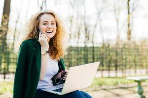 Attractive woman with trendy hairdo sitting in park communicating over her cell phone using modern laptop computer having broad smile while looking aside. People, technology, communication concept photo