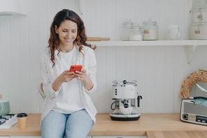 atractiva mujer joven está enviando mensajes de texto en el teléfono inteligente. chica sentada en la encimera de la cocina en casa. foto
