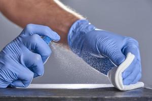Hands in rubber gloves treat the table with a disinfectant spray on a grey background. Disinfection of premises photo