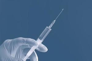 Doctor's white-gloved hand holding a syringe with transparent liquid isolated on a blue background. photo