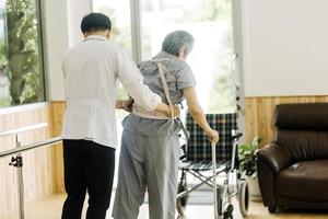 joven fisioterapeuta asiático que trabaja con un anciano usando un andador en el pasillo de un hogar de ancianos foto