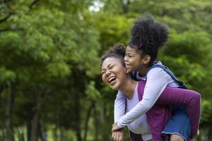 African American mother is playing piggyback riding with her young daughter while having a summer picnic in the public park for wellbeing and happiness concept with copy space photo