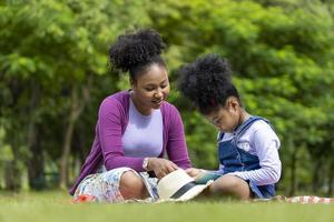 African American mother is teaching her young daughter to read while having a summer picnic in the public park for education and happiness concept photo