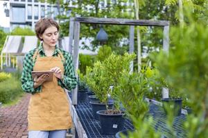 Caucasian gardener holding digital tablet while working in her conifer tree nursery garden center for evergreen and bonsai artist supply photo