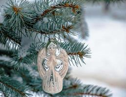 ángel de juguete de navidad cuelga de una rama cubierta de nieve de un árbol de navidad sobre un fondo festivo de nieve blanca bokeh con espacio para copiar. foto