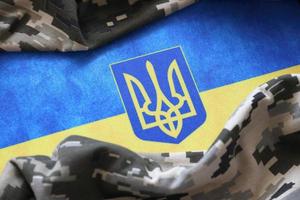 Ukrainian flag and coat of arms with fabric with texture of pixeled camouflage. Cloth with camo pattern in grey, brown and green pixel shapes with Ukrainian trident sign photo