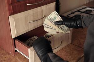 Robber in black outfit and gloves see in opened shelf in kitchen. The thief takes out the US dollar bills from a shelf photo