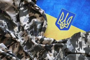 Ukrainian flag and coat of arms with fabric with texture of pixeled camouflage. Cloth with camo pattern in grey, brown and green pixel shapes with Ukrainian trident sign photo