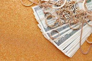 Many expensive golden jewerly rings, earrings and necklaces with big amount of US dollar bills on luxury glitter golden background surface. Pawnshop or jewerly shop photo