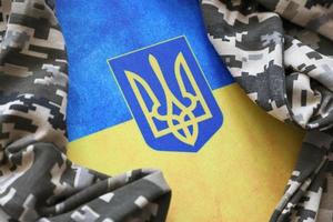 Ukrainian flag and coat of arms with fabric with texture of pixeled camouflage. Cloth with camo pattern in grey, brown and green pixel shapes with Ukrainian trident sign photo