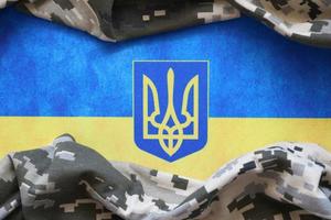 Ukrainian flag and coat of arms with fabric with texture of pixeled camouflage. Cloth with camo pattern in grey, brown and green pixel shapes with Ukrainian trident sign photo