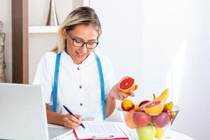 Portrait of young smiling female nutritionist in the consultation room. Nutritionist desk with healthy fruit, juice and measuring tape. Dietitian working on diet plan. photo