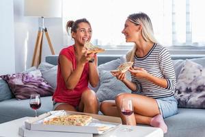 Happy young female friends eating pizza with wine on sofa at home. Happy smiling young women friends eating pizza and drinking wine at home photo