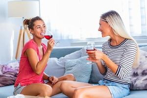 Two attractive girls, cheerful best friends having fun and drinking red wine at home. Two glasses of white wine in hands. Girls having fun time together gossiping photo