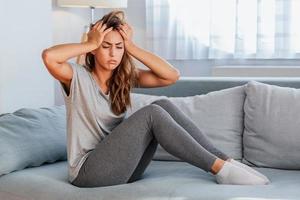 Portrait of an attractive woman sitting on a sofa at home with a headache, feeling pain and with an expression of being unwell. Upset depressed woman lying on couch feeling strong headache migraine. photo