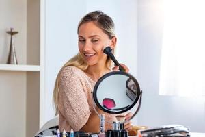 mujer de belleza al maquillarse. hermosa chica mirándose en el espejo y aplicando cosméticos con un cepillo grande. la chica se sonroja en los pómulos. polvo, colorete foto