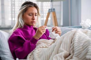 Sickness, seasonal virus problem concept. Woman being sick having flu lying on sofa looking at temperature on thermometer. Sick woman lying in bed with high fever. Cold flu and migraine. photo