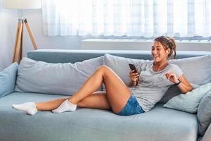 relájate al ritmo de mi música. atractiva mujer joven con auriculares y teléfono inteligente sentado en el sofá en casa. hermosa chica en denim y auriculares está escuchando música usando un teléfono inteligente foto