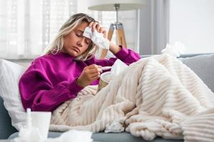 enfermedad, concepto de problema de virus estacional. mujer enferma con gripe tendida en el sofá mirando la temperatura en el termómetro. mujer enferma acostada en la cama con fiebre alta. gripe fría y migraña. foto