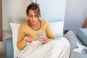 Concept of suffering from terrible pain. Beautiful, sad, upset, unhappy, troubled, weak woman with blonde hair, wearing casual clothes is sitting on a sofa and taking a pill photo