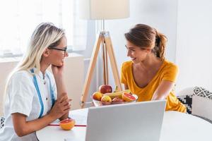 médico nutricionista, dietista y paciente femenino en consulta en el consultorio. joven nutricionista mujer sonriente en la sala de consulta. escritorio nutricionista con fruta saludable y cinta métrica. foto