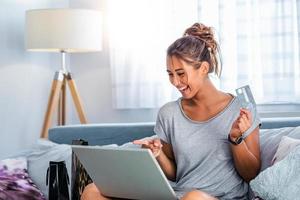 mujer joven en el sofá de compras en línea con tarjeta de débito. hermosa chica usando una computadora portátil para comprar en línea en casa foto