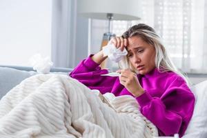 Sickness, seasonal virus problem concept. Woman being sick having flu lying on sofa looking at temperature on thermometer. Sick woman lying in bed with high fever. Cold flu and migraine. photo