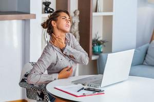 Portrait of young stressed woman sitting at home office desk in front of laptop, touching aching back with pained expression, suffering from backache after working on pc photo