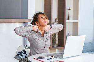 Businesswoman feeling pain in neck after sitting at the table with laptop. Tired female suffering of office syndrome because of long hours computer work. Pretty girl massaging her tense neck muscles photo