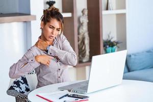 retrato de una joven mujer estresada sentada en el escritorio de la oficina en casa frente a una laptop, tocándose la espalda con expresión de dolor, sufriendo de dolor de espalda después de trabajar en la pc foto