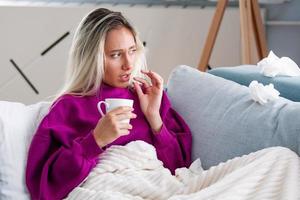 Concept of suffering from terrible pain. Beautiful, sad, upset, unhappy, troubled, weak woman with blonde hair, wearing casual clothes is sitting on a sofa and taking a pill photo