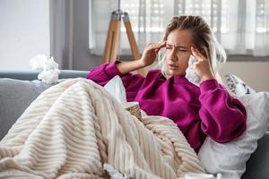 Sick woman with headache sitting under the blanket. Sick woman with seasonal infections, flu, allergy lying in bed. Sick woman covered with a blanket lying in bed with high fever and a flu, resting. photo