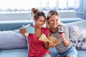 Friends eating pizza and smiling for selfie. They are sharing pizza and making selfie photo on mobile smart phone. They are having party at home, eating pizza and having fun.