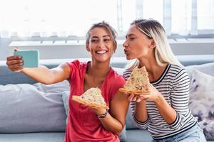 Friends eating pizza and smiling for selfie. They are sharing pizza and making selfie photo on mobile smart phone. They are having party at home, eating pizza and having fun.