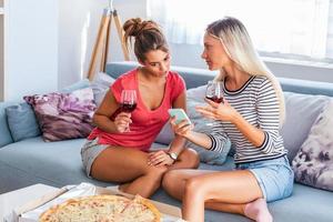 Two female friends gossiping with glasses red wine and pizzas, Showing images to her friend on her social network on the mobile phone photo
