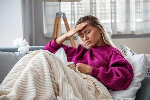 Sick woman with headache sitting under the blanket. Sick woman with seasonal infections, flu, allergy lying in bed. Sick woman covered with a blanket lying in bed with high fever and a flu, resting. photo