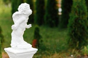 estatua de ángel bebé con alas contra jardín verde. el lindo angelito cupido escultura estatua de pie al aire libre foto