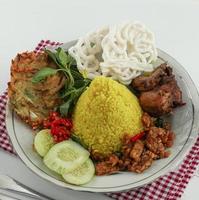 Nasi kuning or yellow rice in cone shape,Indonesian festive rice dish presentation with some condiments, like chicken, egg, tempe,cucumber,chili wih red napkin photo