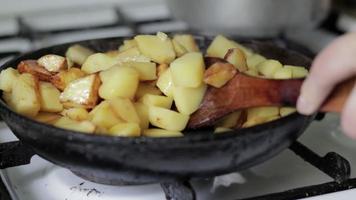 a mão de uma mulher mexe batatas fritas com uma espátula de madeira em uma frigideira. o processo de cozinhar batatas frescas em uma panela de ferro fundido com óleo de girassol. uma frigideira cheia de batatas fritas douradas. video