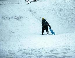 niño pequeño trineo cuesta abajo en invierno. el niño cae en la nieve. juego al aire libre para niños. trineos para niños en un parque cubierto de nieve. foto