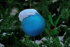 A blue Christmas ball hangs on a snow-covered branch of a Christmas tree on a festive background of white snow and snowy bokeh with copy space. photo