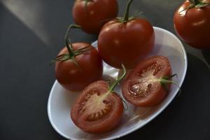 Beautiful fruits of a whole and cut tomato on a white plate side view. Delicious tomatoes still life in the rays of the evening sun on a black background. photo