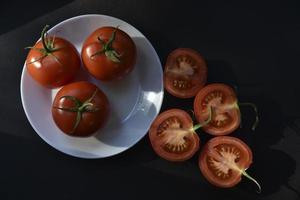 Beautiful fruits of a whole and cut tomato on a white plate side view. Delicious tomatoes still life in the rays of the evening sun on a black background. photo