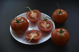 Beautiful fruits of a whole and cut tomato on a white plate side view. Delicious tomatoes still life in the rays of the evening sun on a black background. photo