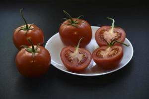 Beautiful fruits of a whole and cut tomato on a white plate side view. Delicious tomatoes still life in the rays of the evening sun on a black background. photo