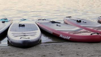 Empty SUP boards sway on the waves on a calm river or lake. Water sports. Lots of boards at the rental shop, the sun reflecting off the ripples of the water. Panning. Ukraine, Kyiv - August 07, 2022. video