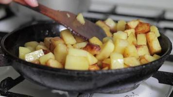 A woman's hand stirs fried potatoes with a wooden spatula in a frying pan. The process of cooking fresh potatoes in a cast iron pan with sunflower oil. A frying pan filled with golden fried potatoes.
